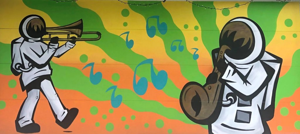 Two astronauts in space suits. One walking and playing a trombone, the other playing a saxophone against a green, orange, and yellow wavy radial striped background. There are lime green polkadots and music notes floating around in the background