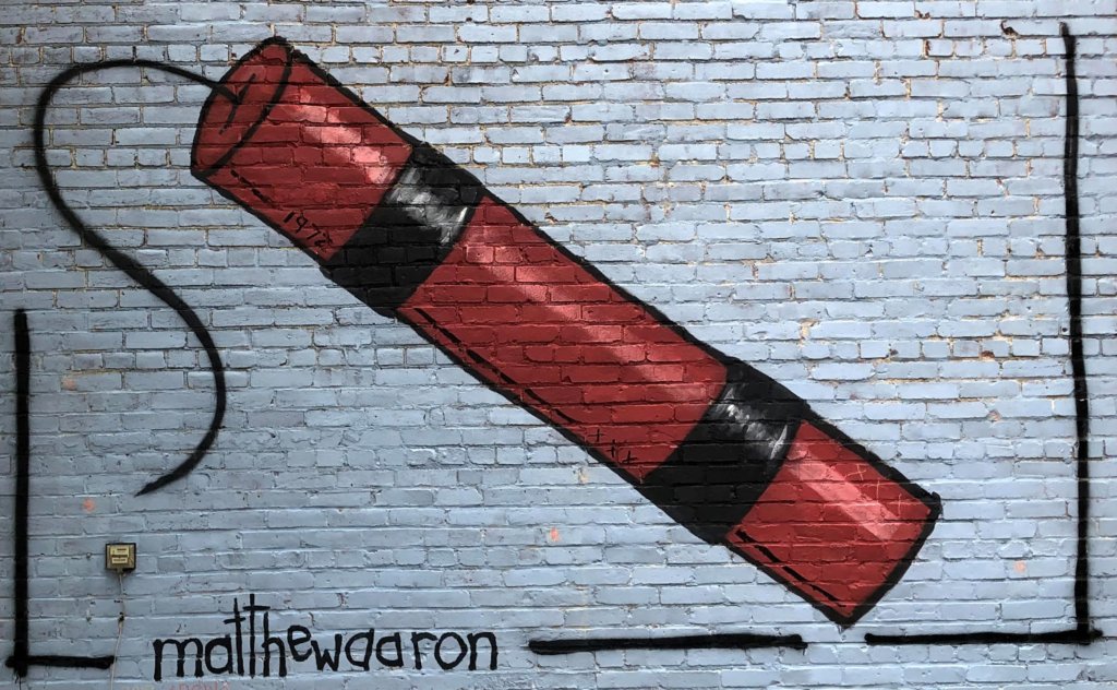 Blue rectangle with single stick of bright red dynamite diagonally across it. Framed with a single black spray paint line and signed matthewaaron