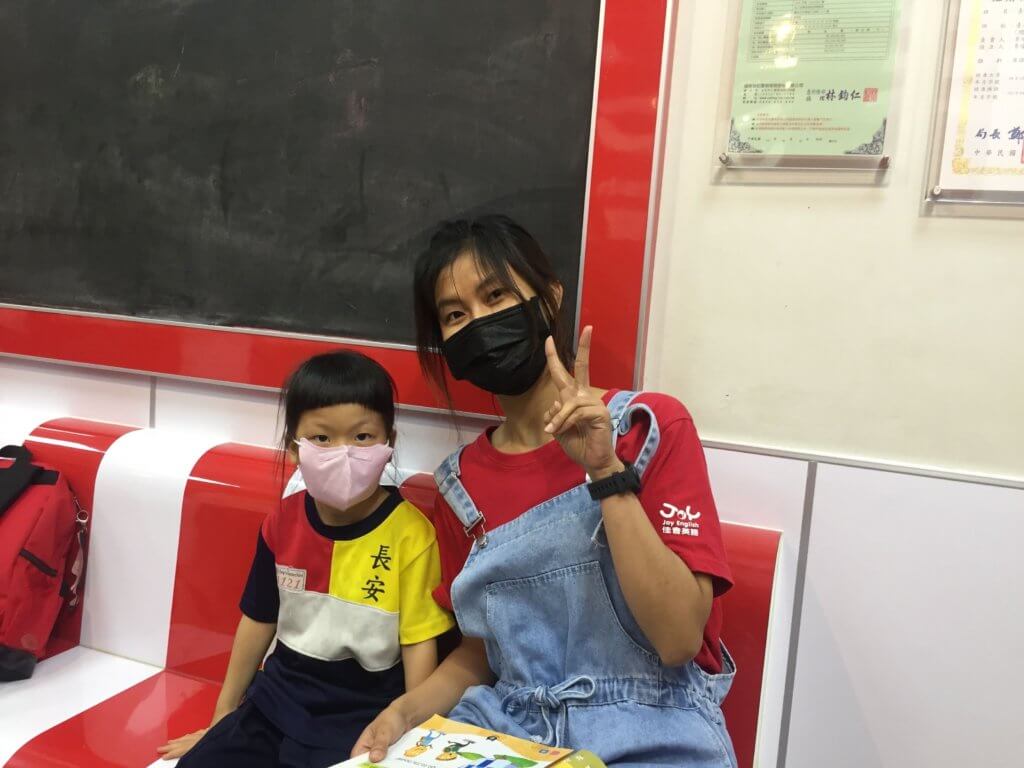 Asian woman poses with elementary aged Taiwanese student. She is wearing a red t-shirt and blue overalls. The kid is a young girl wearing a red, yellow and Black shirt